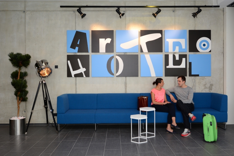 Zwei Gäste auf einem gemütlichen blauen Sofa in der Hotellobby unter einem Bilderarrangement, das die Worte 'Arte Hotel' ergibt
