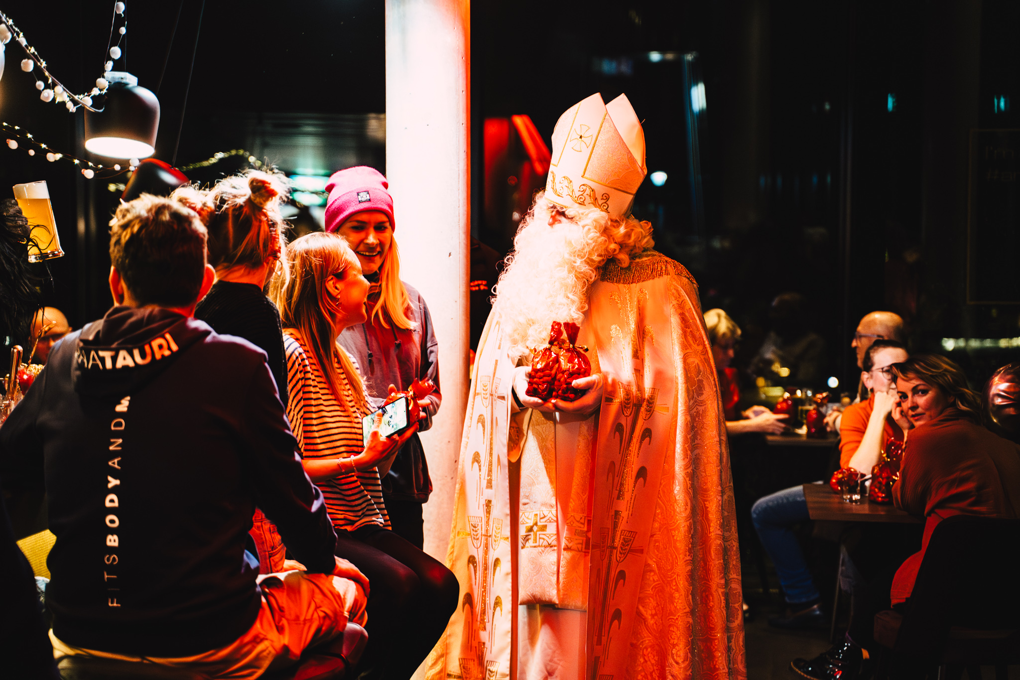 Nikolaus in weiß-goldener Robe überreicht ein kleines Geschenk an lachende Gäste in einer Rooftop Bar.