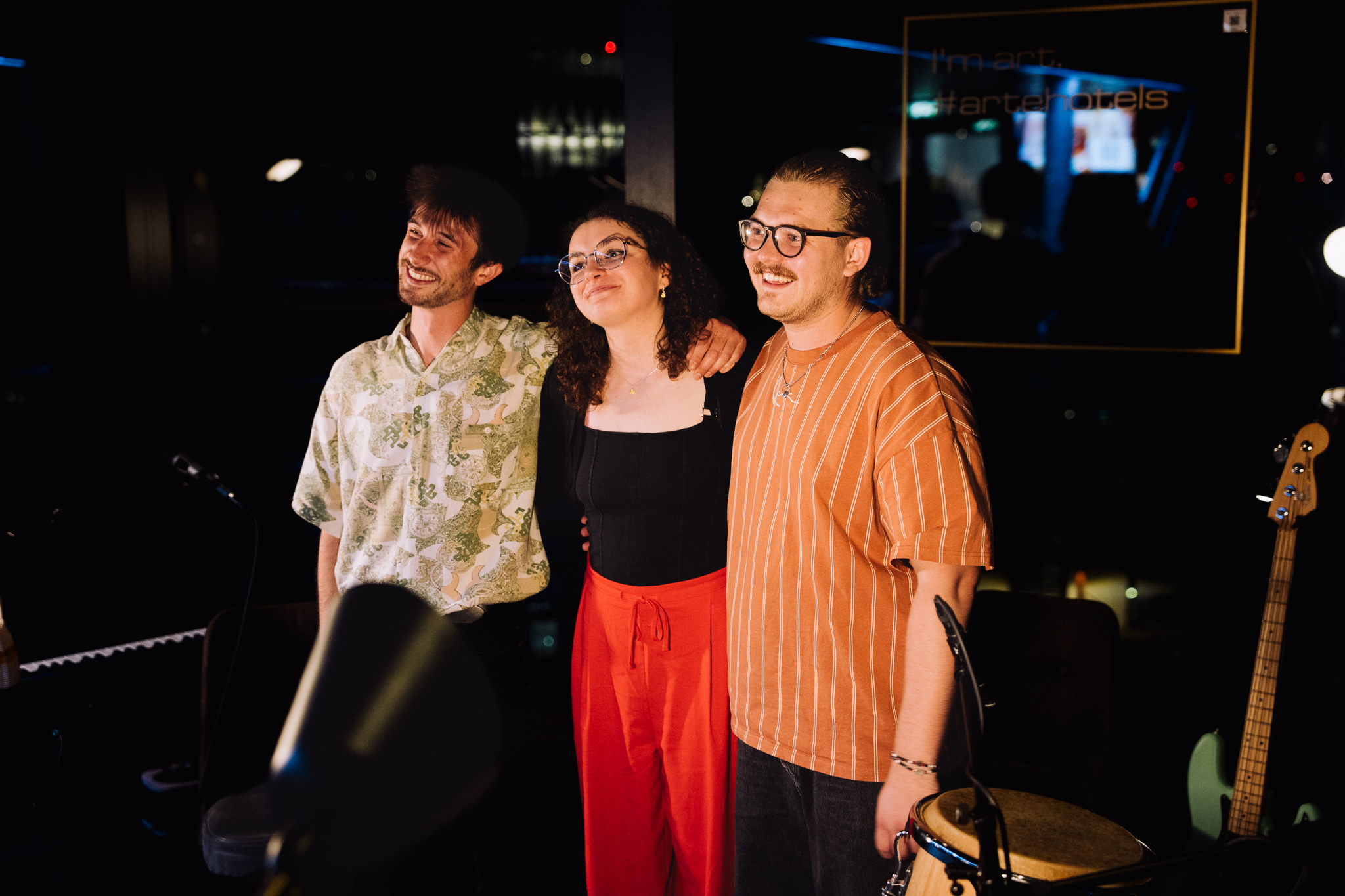 Three smiling musicians pose after a live performance in a warmly lit venue, surrounded by microphones and musical instruments.
