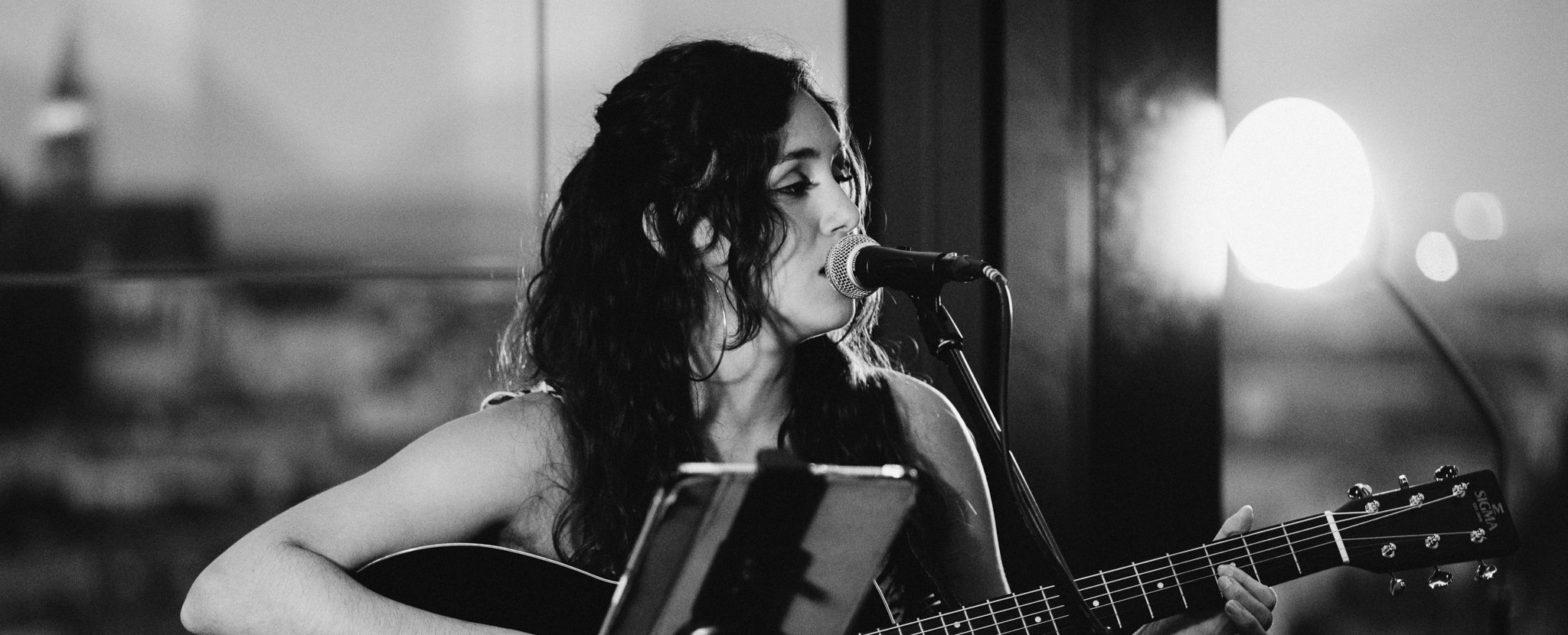 Kati Castillo performing live with a black acoustic guitar and microphone at the hu:goes14 rooftop bar in a black-and-white photo.