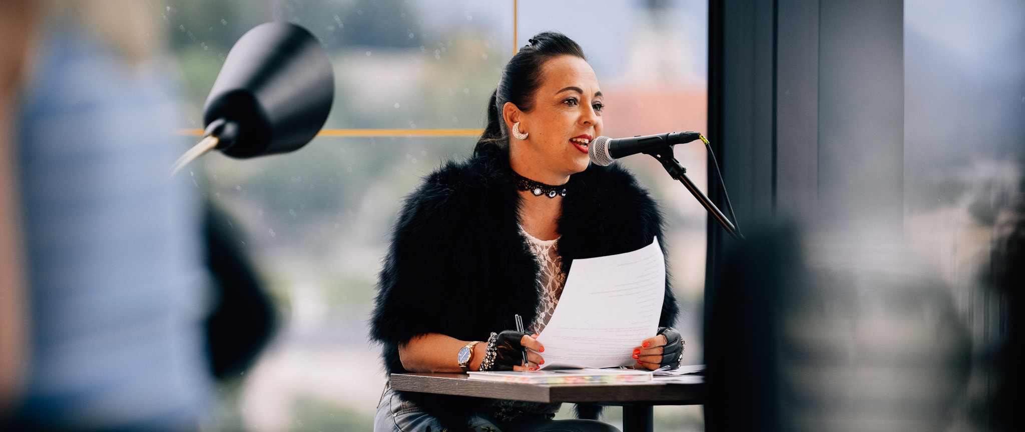 Jasmine Wallner sits at a table during her reading at arte Hotel Salzburg and speaks into a microphone.