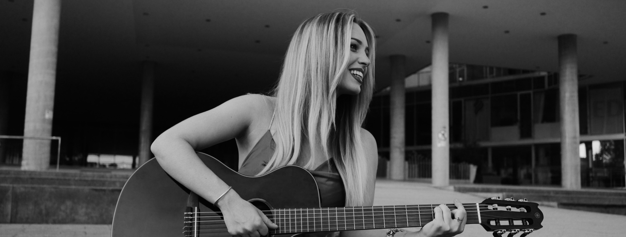 Junge Musikerin sitzt mit Akustikgitarre vor einem modernen Gebäude und lächelt während des Spielens.