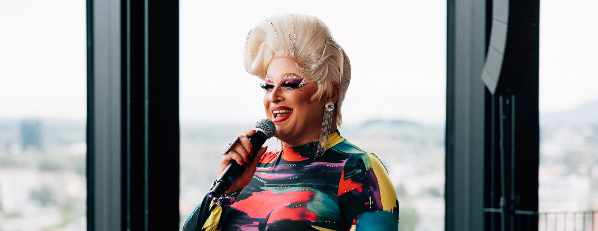 Drag-Queen mit Mikrofon bei den Brunching Divas im hu:goes14 Rooftop-Bar des arte Hotel Salzburg, vor großen Fenstern mit Blick über die Stadt.