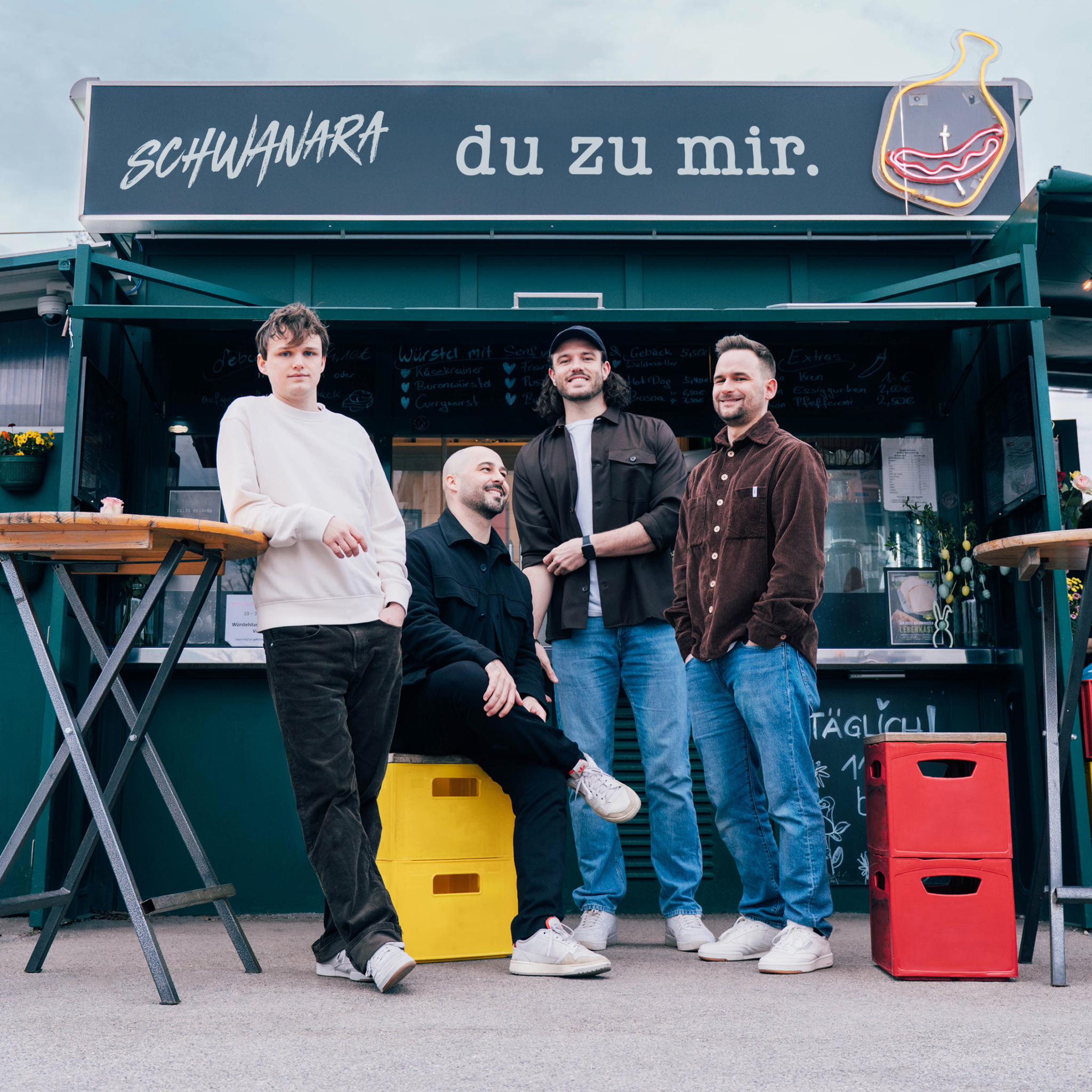 Die vier Mitglieder der österreichischen Austropop-Band Schwanara posieren vor einem dunkelgrünen Imbissstand mit der Aufschrift „du zu mir.“ Drei Musiker stehen, während einer auf gelben Getränkekisten sitzt. Über ihnen sind der Bandname und ein beleuchtetes Neonschild zu sehen.