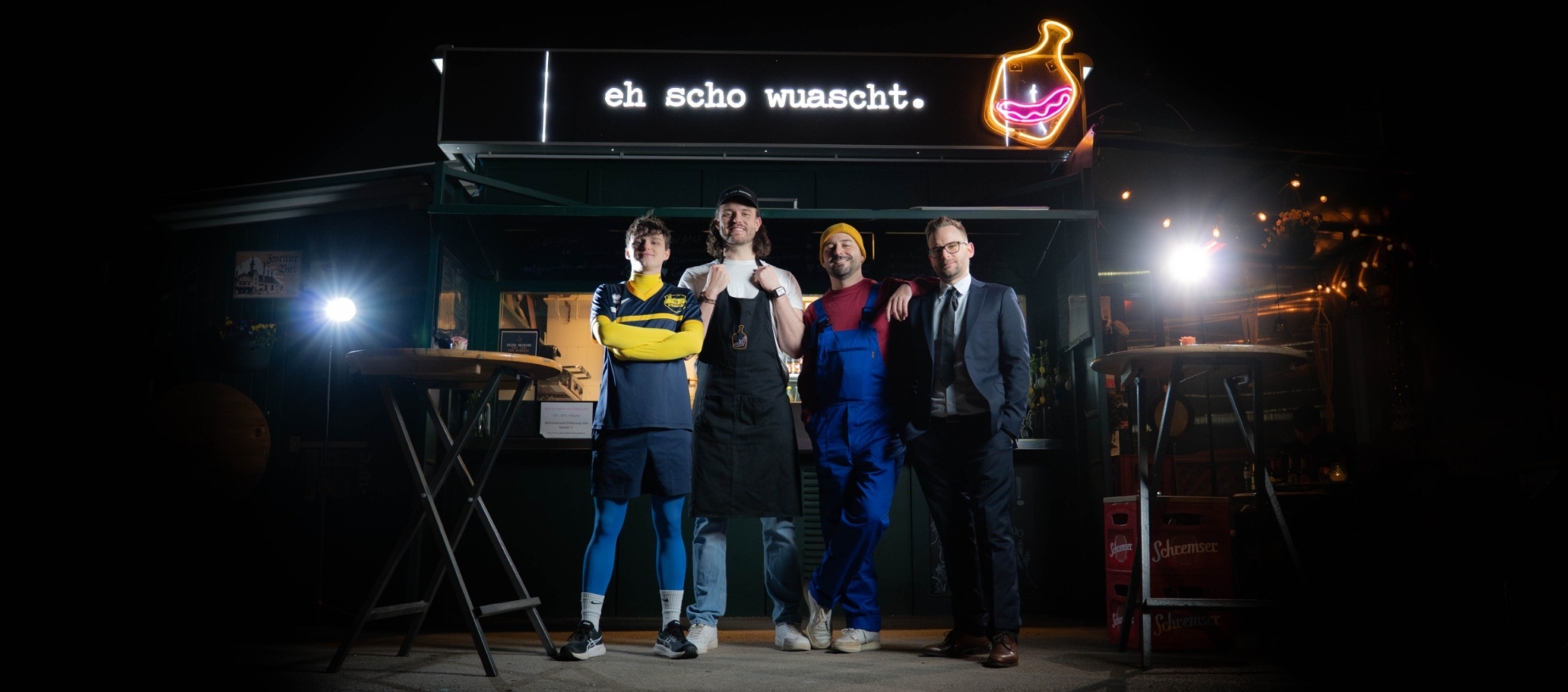 The four members of Austrian band Schwanara pose at night in front of an illuminated food stand featuring the words “eh scho wuascht.” The musicians wear a variety of playful outfits.