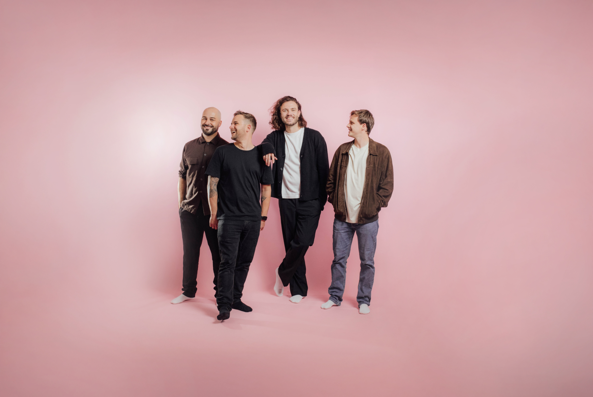 The four members of Austrian pop band Schwanara stand in casual clothing in front of a large blue-grey metal facade. The group portrait captures the musicians in relaxed, individual poses.